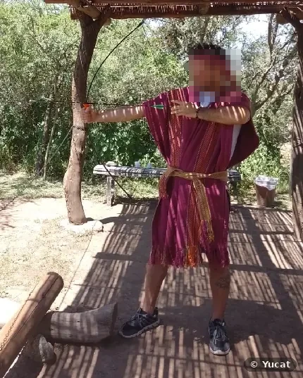 Un hombre con poncho bordó y vincha tensa un arco bajo un quincho, entre árboles.