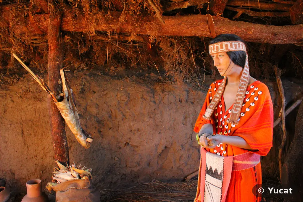 Interior de una casa-pozo comechingona: una escultura con vincha tejida junto a un arco y un carcaj colgados, y vasijas de barro con choclos.