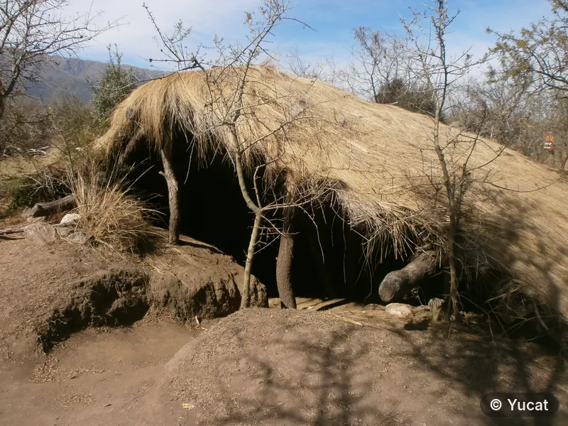 Vista exterior de una casa-pozo con techo de paja, con las sierras de fondo.