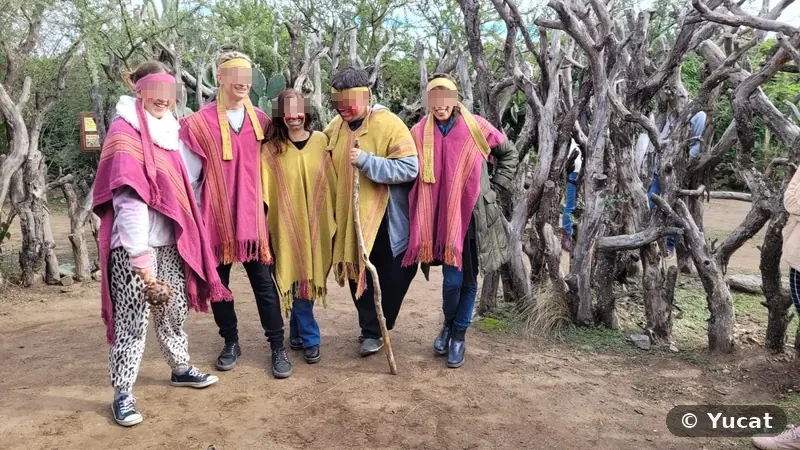 Cinco visitantes con ponchos rosas y amarillos, vinchas y pintura facial, abrazados en el sendero junto a un cerco de troncos retorcidos.