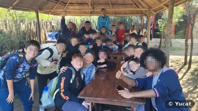 Un grupo escolar posa sonriente con sus acompañantes alrededor de las mesas del área de descanso, bajo un techo de paja.