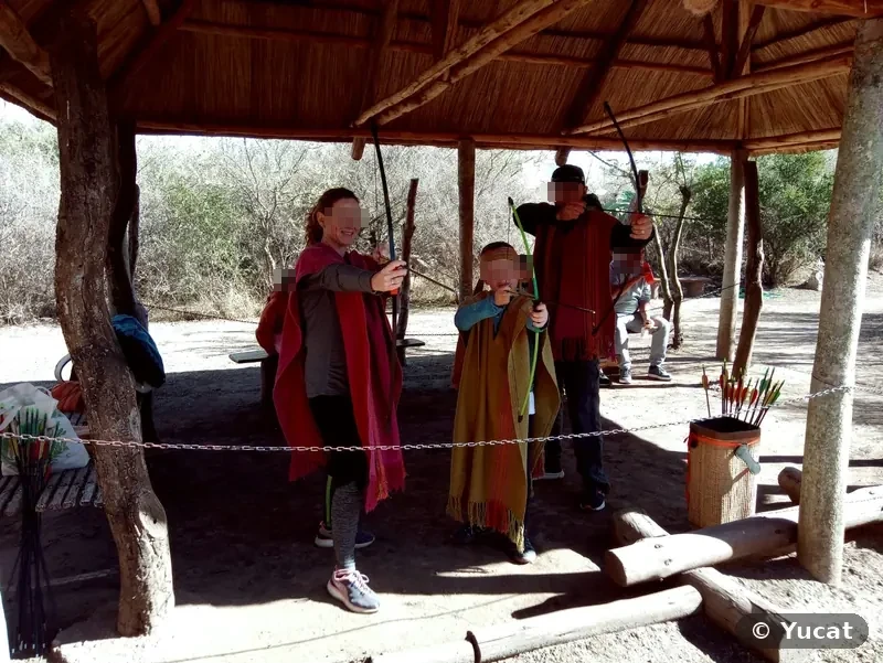Una mujer, un chico y un hombre con ponchos practican tiro con arco bajo un quincho de paja.