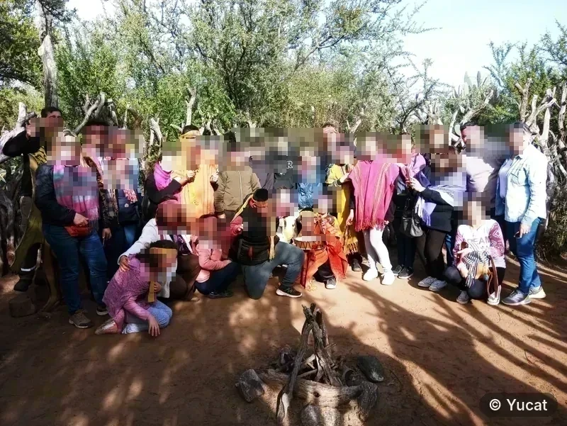 Grupo de visitantes con vinchas y pañuelos tejidos, sonriendo alrededor de un fogón de piedras y leños.