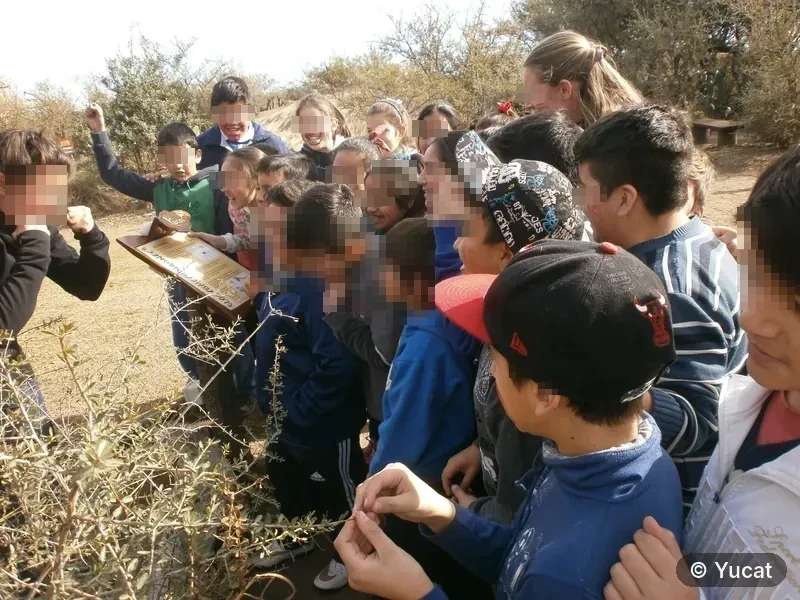 Chicos reunidos alrededor de un cartel de madera durante una visita, en un sendero con arbustos.