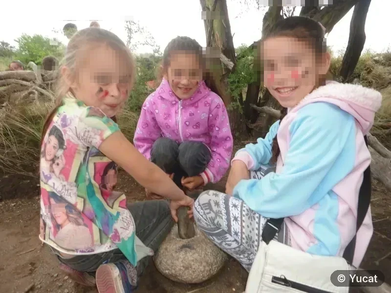 Tres chicas con la cara pintada moliendo granos en un mortero de piedra, junto a un árbol.
