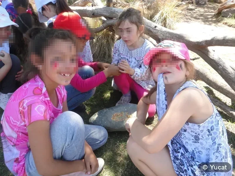 Un grupo de chicas con la cara pintada, sentadas junto a un mortero de piedra bajo un tronco caído.