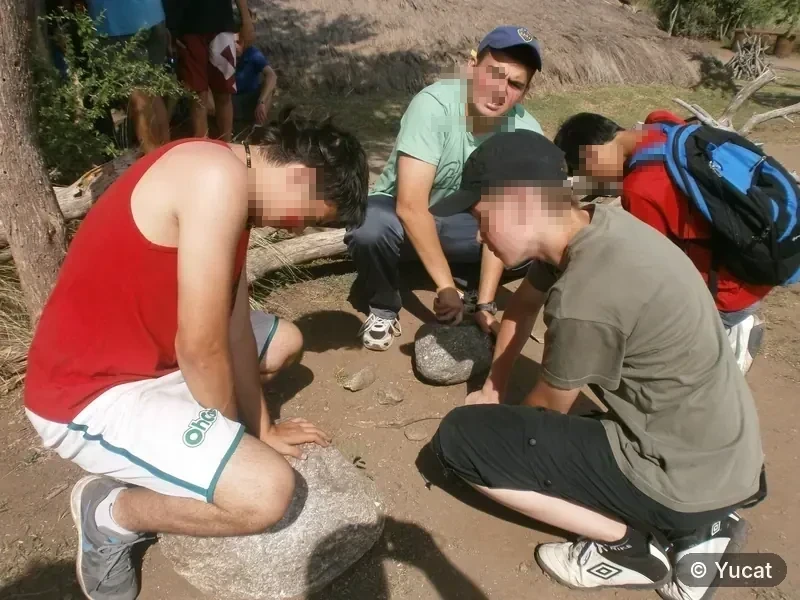Un grupo de jóvenes moliendo granos en morteros de piedra junto a una casa-pozo.