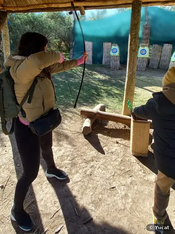 Una visitante, de espaldas, tensa un arco bajo un techo de paja, con los blancos al fondo; a su lado, un chico junto a la caja de flechas.