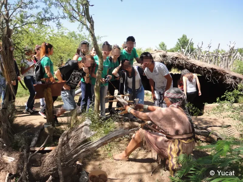 Un grupo de visitantes observa una demostración de telar de cintura junto a una casa-pozo.